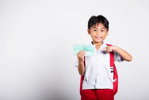 Asian student kid boy wearing student thai uniform and pointing finger to protect mask before to go to school in studio shot isolated on white background, preschool, new normal back to school
