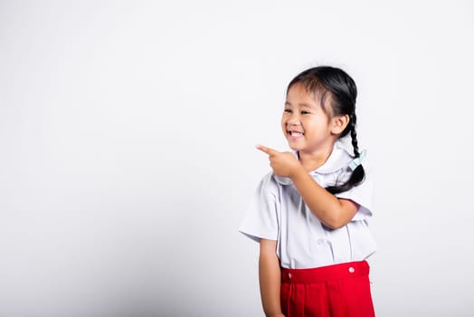 Asian toddler smiling happy wear student thai uniform red skirt keeps pointing finger at copy space in studio shot isolated on white background, Portrait little children girl preschool, Back to school
