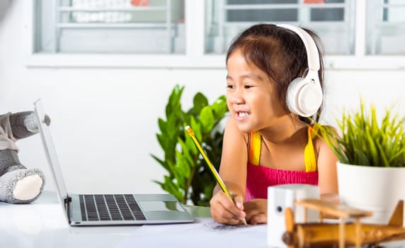 Little kid preschool wear headphone sit at desk use laptop computer and communicates on Internet online video call with teacher, Asian child girl study video conference distant education at home