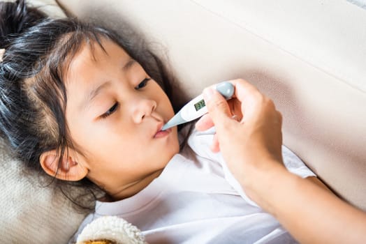Sick kid. Mother checking temperature of her sick daughter with thermometer in mouth, child laying in bed taking measuring her temperature for fever and illness