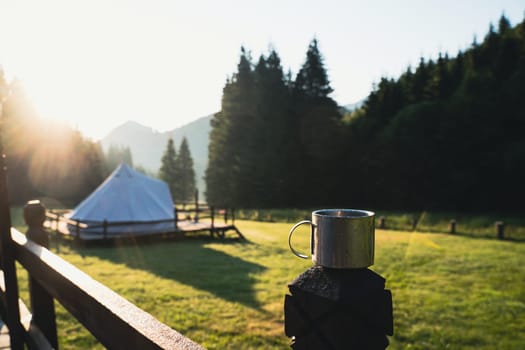 closeup front view of metallic coffee cup in the morning with glamping tent camping ground in the background in a green mountain meadow surrounded by fir tree forest