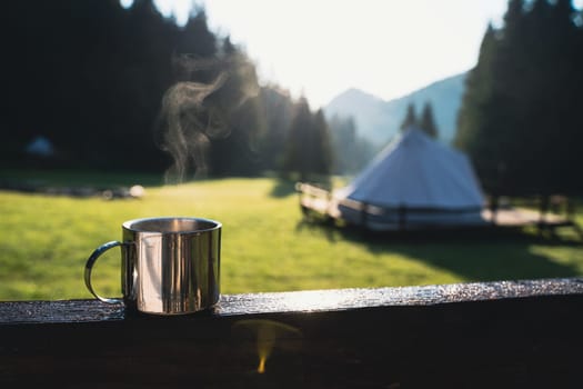 closeup front view of a steaming metallic cup with warm coffee or tea in the morning with glamping camping ground in the background