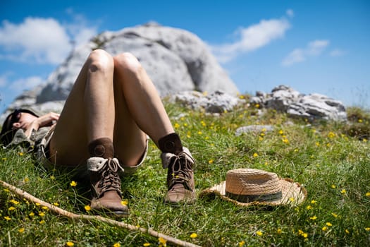 young Caucasian woman lying down and taking a rest in the warm sun near a wooden trekking stick and wicker hat in the grass on a mountain peak with blue sky and clouds above
