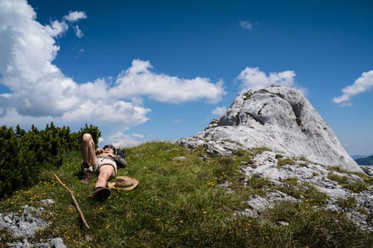 young Caucasian woman lying down and taking a rest in the grass on a mountain peak with blue sky and clouds above