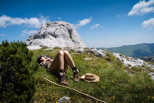 young Caucasian woman lying down and taking a rest in the warm sun near a wooden trekking stick and wicker hat in the grass on a mountain peak with blue sky and clouds above