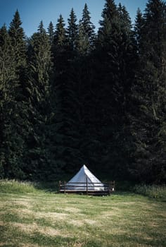 front view of secluded glamping camping tent with white cotton fabric on wooden deck in green meadow surrounded by fir tree forest