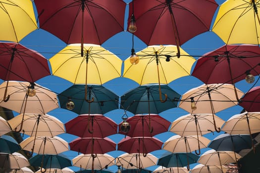 front view closeup detail of colorful umbrellas and Edison light bulbs hanging out above a city street against a blue sky