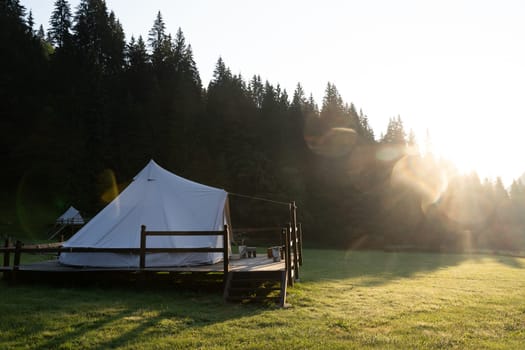 side view of white glamping tent on wooden deck in green meadow surrounded by fir tree forest at sunrise