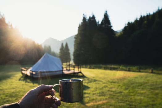 closeup front view hand holding a metallic coffee cup in the morning with glamping tent camping ground in the background in a green mountain meadow surrounded by fir tree forest