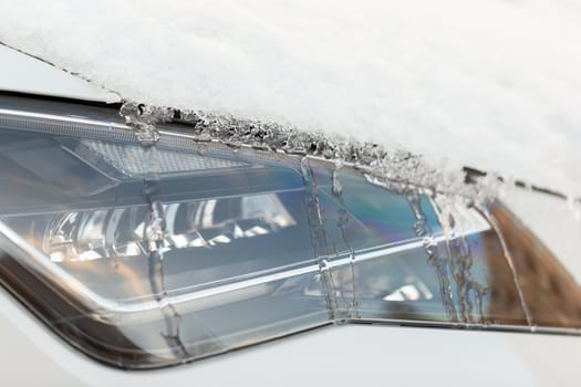 Close up of a headlight of a car with ice melting and water running down the bumper. Detailed view of a vehicle part covered in snow. Concept of transportation problems in wintertime.