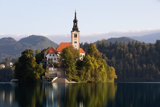 Church in Bled lake illuminated from side view at sunrise with clear water surface reflecting green nature around. Catholic building accessible by boat in Europe with sky in background.