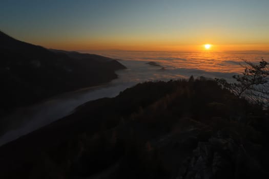 Sun rising above low clouds in mountain with dark hills in background. Nature scenery early in the morning with illuminated sky and valley still hidden in shadow.