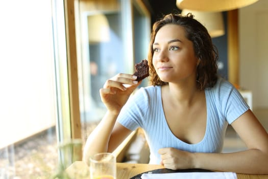 Woman holding and eating a chocolate cupcake sitting in a restaurant looking through a window