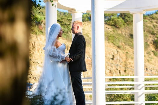 Young beautiful interracial newlyweds lovingly look at each other against the background of white columns