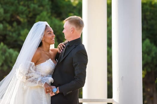 Half-length portrait of mixed-racial newlyweds on a walk against the background of columns and greenery