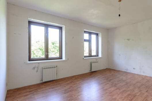 The interior of an empty room during renovation, there are two large windows in the room, radiators under them