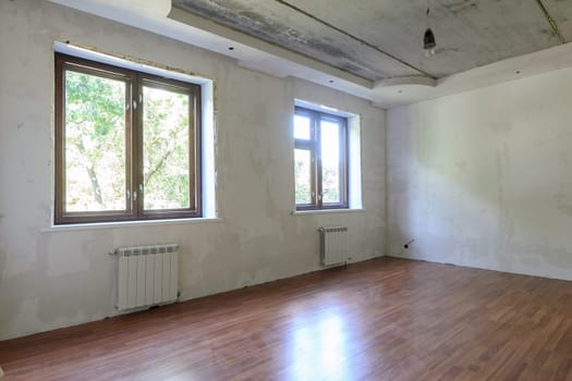 The interior of an empty room during renovation, there are two large windows in the room, heating radiators are located under the windows