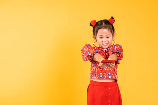 Chinese New Year. Happy Asian Chinese little girl smile wear red cheongsam handed angpao or red packet monetary gift, Portrait children in traditional dress, studio short isolated on yellow background