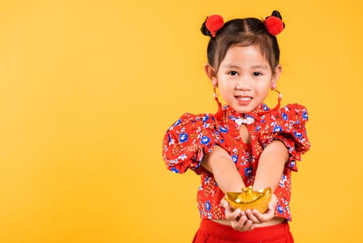 Chinese New Year. Happy Asian Chinese little girl smile wearing red cheongsam holding gold ingot, Portrait children in traditional dress hold golden bar, studio short isolated on yellow background