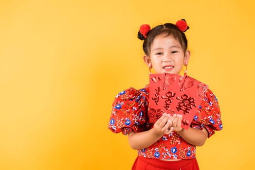 Chinese New Year. Happy Asian Chinese little girl smile wear red cheongsam qipao hold angpao red packet monetary gift, Portrait child in traditional dress, studio short isolated on yellow background