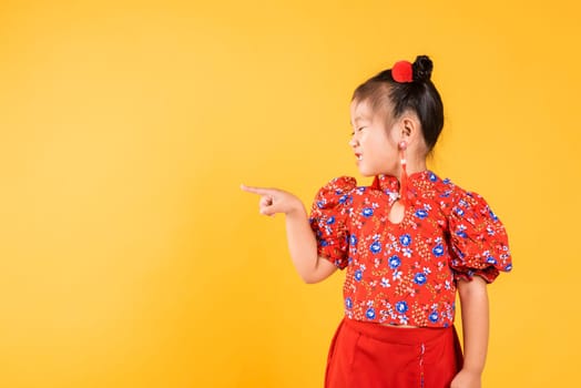 Happy Asian Chinese little girl wear red cheongsam qipao pointing finger to space, Portrait children in traditional dress point to empty, Chinese New Year, studio short isolated on yellow background