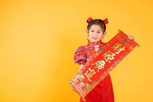 Chinese New Year. Happy Asian Chinese little child girl smile wearing red cheongsam qipao Spring festival couplets (Character "FU"" means fortune, blessing), studio short isolated on yellow background"