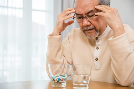 Asian elderly man sick have headache looking pill glass on at home, senior old man grey haired sad sitting in chair hand hold head with many medicines and pills for taking, Elderly healthcare