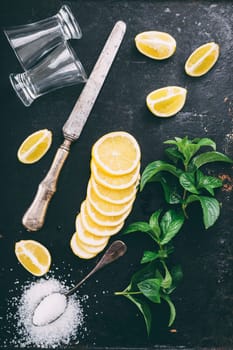 Top view of cocktail ingredients preparation on black rustic background. Tequila shot glasses, lemon slice, salt and mint on grunge dark table form above. Cocktail recipe making.