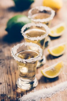 Tree golden mexican tequila shot cocktail with lime slices and salt in row on bar counter with ingredients. Hard liquor glass and lemon on rustic wooden restaurant table. Retro style filter.