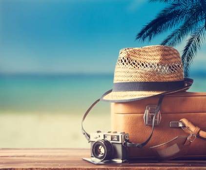 Vintage suitcase, hipster hat, photo camera and passport on wooden dack. Tropical sea, beach and palm three in background. Summer holiday traveling design concept.