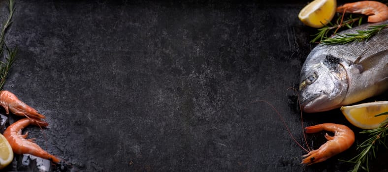 Top view of fresh sea food ingredients on grunge textured black background design. Shrimps, bream sea fish, herbs, lemon and ice on rustic dark slate with copyspace.
