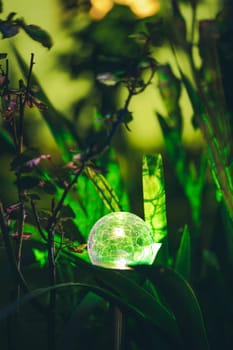 Night View Of Flowerbed Illuminated By Energy-Saving Solar Powered Colorful Multi-colored Lantern On Yard. Beautiful Small Garden With Green Light, Lamp In Flower Bed. Garden Design.