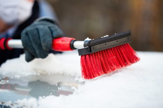 Man in gloves removing snow from a roof of a car with red brush. Vehicle maintenance in freezing conditions. Human cleansing automobile of ice in detailed view.