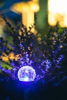Night View Of Flowerbed Illuminated By Energy-Saving Solar Powered Colorful Multi-colored Lantern On Yard. Beautiful Small Garden With Blue Light, Lamp In Flower Bed. Garden Design.
