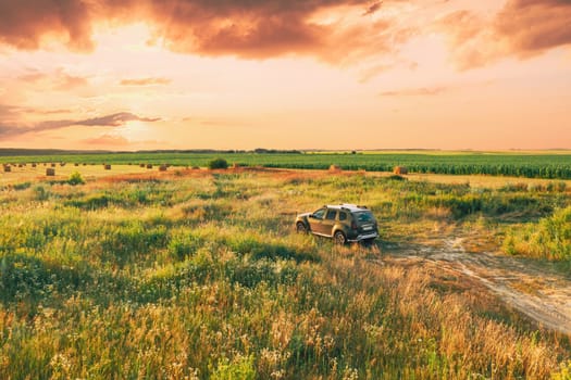 Elevated View Of Car SUV Parked Near Countryside Road In Summer Field Rural Landscape. Altered Sunset Sunrise Sky Above Meadow Rural Landscape.