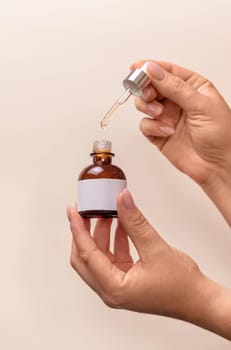 Hands holding pipette and opened dropper bottle with blank label over light beige, close up, mockup. Natural cosmetic, serum or essence. Everyday woman skincare routine.