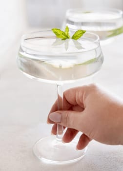 Female hand holding transparent cocktail in a coupe glass decorated with mint leaves close up. Tropical drink at the bar. Summer vacation mood. Analcolic mocktail drink