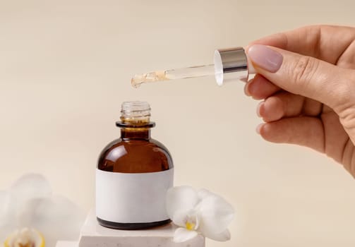 Hand holding pipette over opened cosmetic dropper bottle near white orchid flowers and stones on light beige, close up, mockup. Natural skincare product, serum or essential oil.