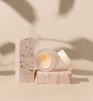 Smass Glass Cosmetic jar on travertine stones on light beige close up, hard shadows. Skincare beauty product package. Exotic natural cosmetics, pastel minimal composition..