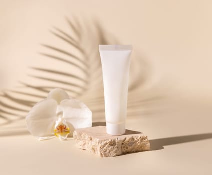 White plastic cream tube on travertine stone near white orchid flower on light beige, Palm leaf hard shadows, close up, mockup. Skincare beauty product. Natural cosmetics. Tropical composition..
