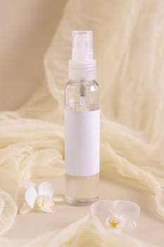 Spray bottle filled with liquid near orchid flowers and tulle on light beige close up. Packaging Mockup. Exotic natural cosmetics, romantic composition, everyday skincare routine..