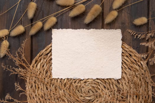 Blank card and envelope on wattled placemat on wood near Hare's Tail grass top view. Boho scene with horizontal paper card. Bohemian or close to nature greeting or Invitation mockup, place for text.