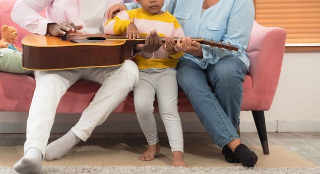 Happy african family with little curly girl sitting relax on pink couch and playing guitar together in living room at home