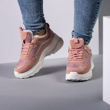 Cropped image of warm pink sneakers with down on female model legs in studio. Sports shoes on feet of unknown young woman who is dressed in jeans and stands on gray floor on background of white cubes.