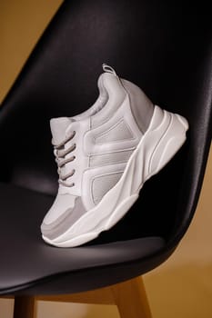 Showing details on sneakers. Close up of one white sneaker standing leaning on back of black chair in studio on beige background. Promotional photo. Side view.
