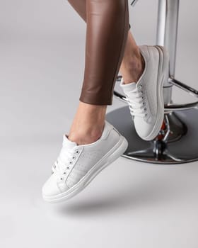 Stylish modern women's sneakers on legs of model sitting on chair in studio on white background. Sneakers are very popular among teenagers or women because of their simple and fashionable appearance.