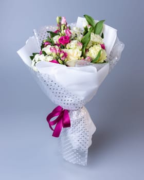 Bouquet of roses, eustoma and irises on a blue background. Flowers are wrapped in white floral paper of two types and tied with a pink ribbon. Concept of floristics and composing greeting bouquets.