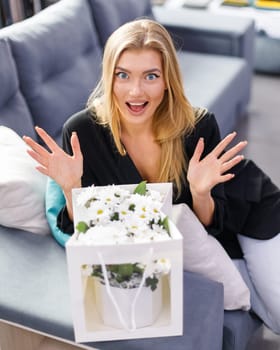 Happy excited woman is pleasantly surprised by bouquet of flowers delivered to her. Beautiful blonde woman exclaims with happy and surprised expression looking at camera while sitting on sofa.