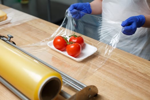 Worker wraps tray with tomatoes with cling film which he pulls from large roll standing on stand on table. Close up of batch of tomatoes for sale, delivery or better storage in refrigerator.