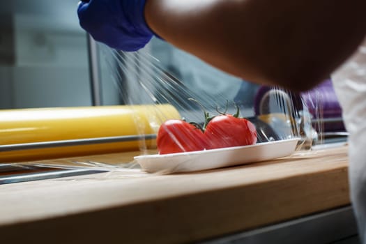 Grocery retailers. Packing of food products. Unknown worker in blue disposable gloves wraps in food transparent film tomatoes lying on white plastic tray. Food delivery concept.